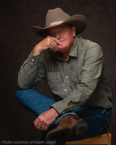 raig Johnson author photo- cowboy hat, blue jeans and gray button-up shirt.